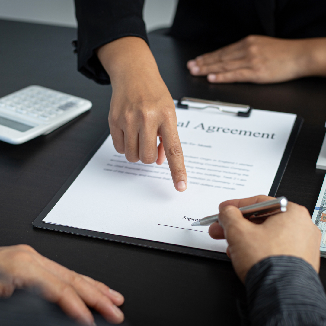 Close-up of a hand pointing to a legal agreement on a clipboard while another hand holds a pen near the signature line, with a calculator and cash nearby.