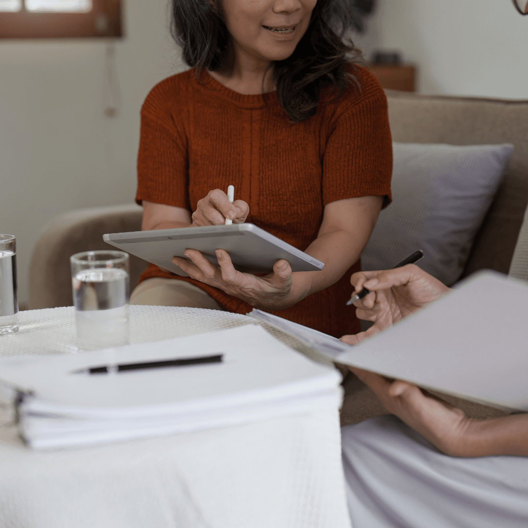 A woman in an orange sweater reviewing documents on a tablet while sitting across from someone holding papers, with water glasses and a stack of paper on the table.