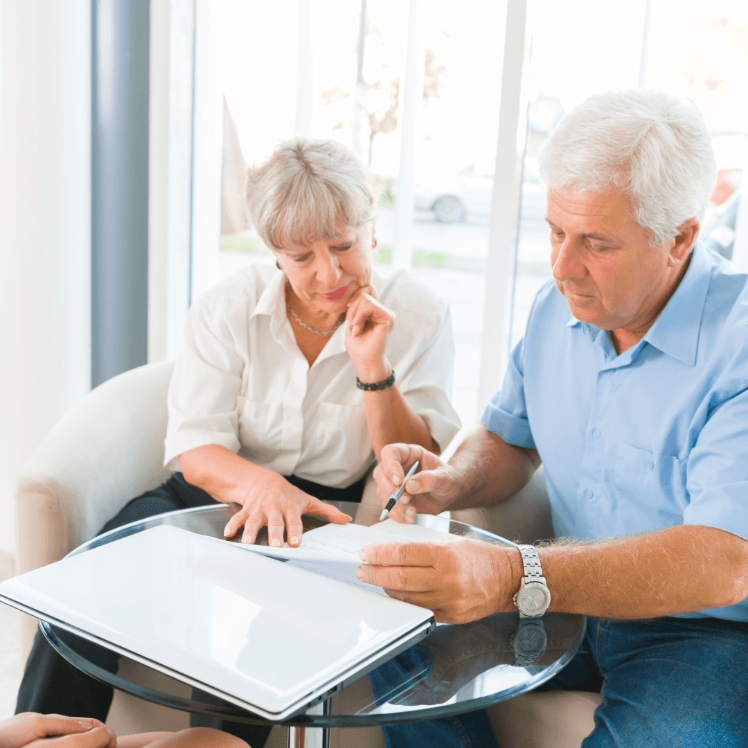 An older couple reviewing and signing a document together at a glass table, with a laptop open beside them.