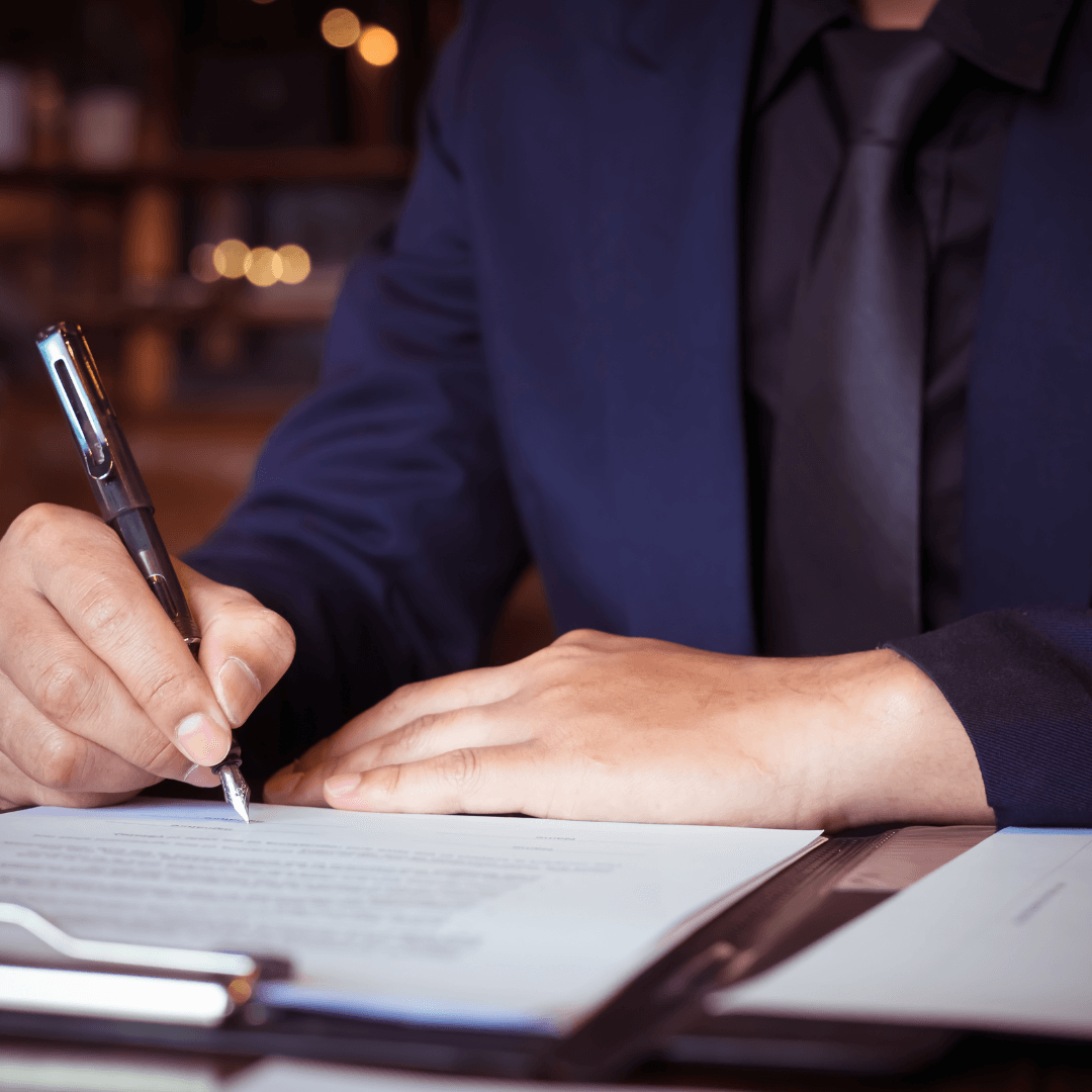 Close-up of a person in a suit signing a document with a fountain pen, papers on a clipboard in the foreground.