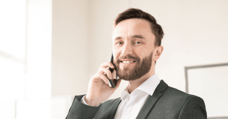 A smiling bearded man in a dark blazer and white open-collared shirt holds a smartphone to his ear in a bright, softly lit room.