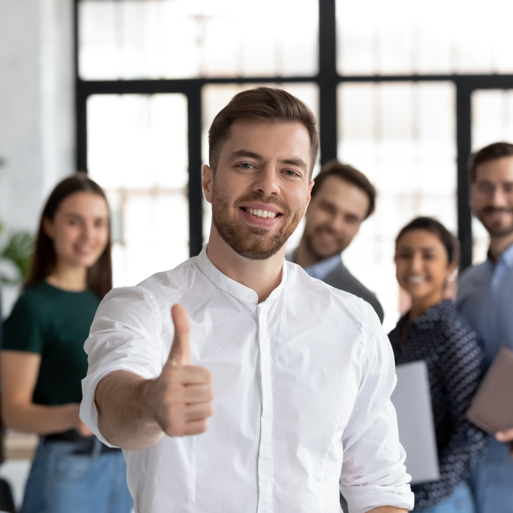 A smiling small business owner gives a thumbs up while standing in front of his happy team in an office setting.