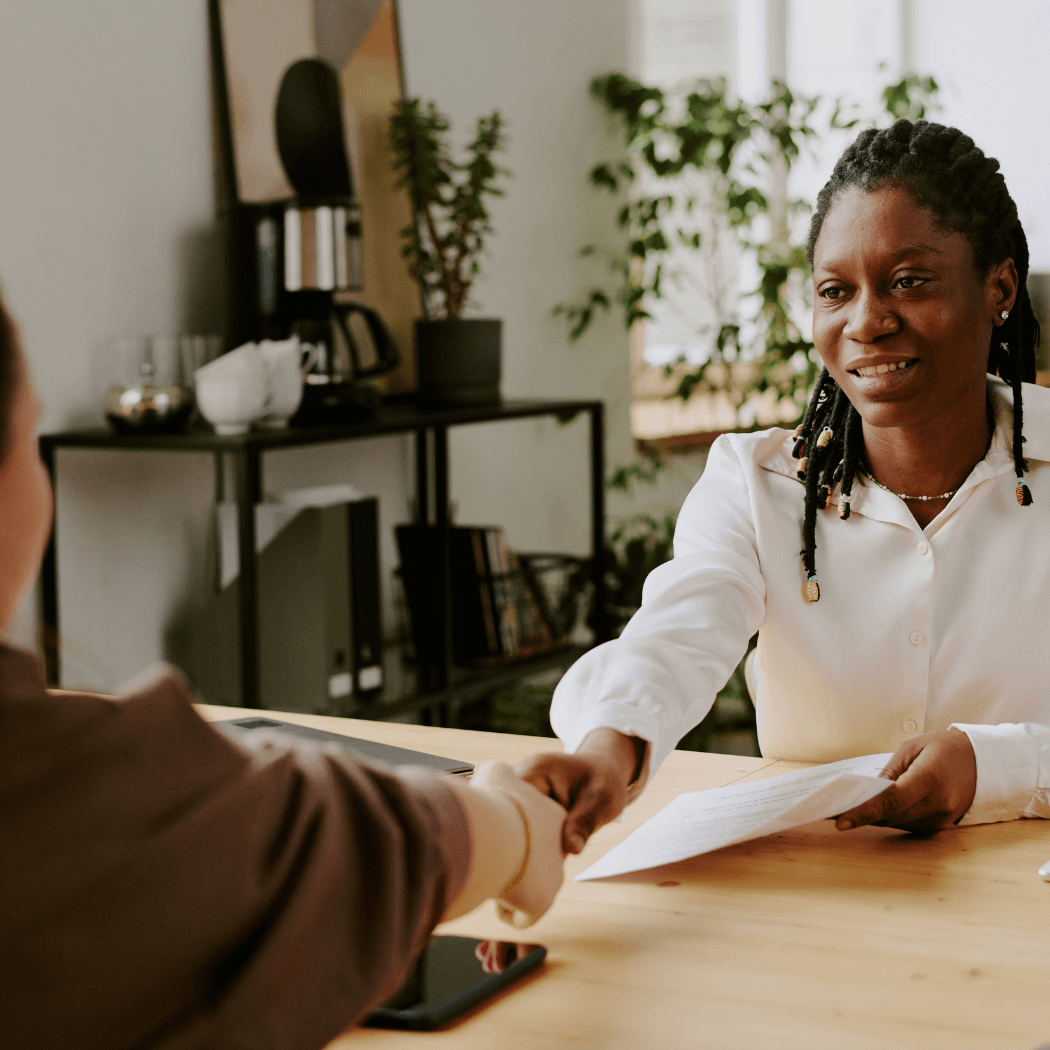 A smiling professional shakes hands with a client across a desk while holding documents during a business meeting.