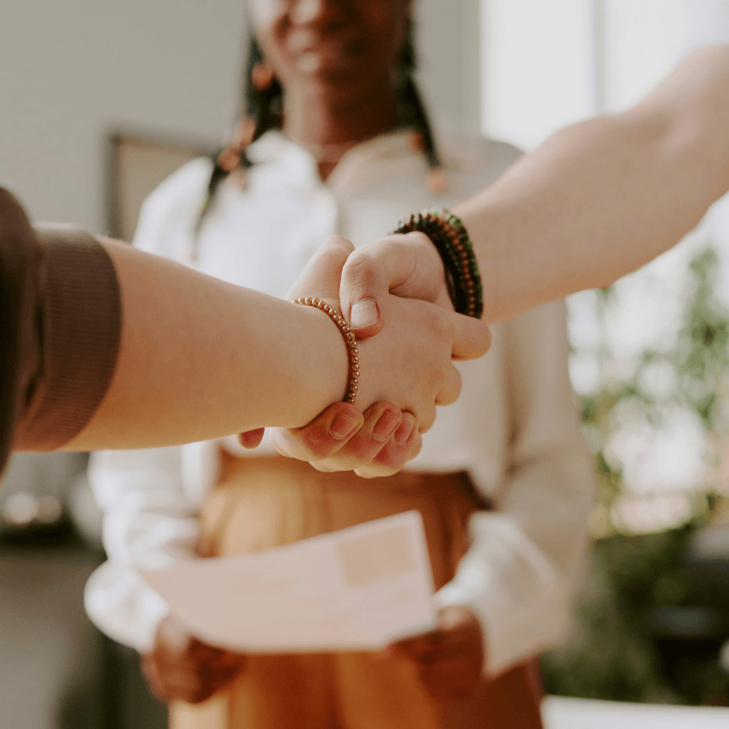 A close-up of two people shaking hands in an office setting, with a third person holding documents in the background.