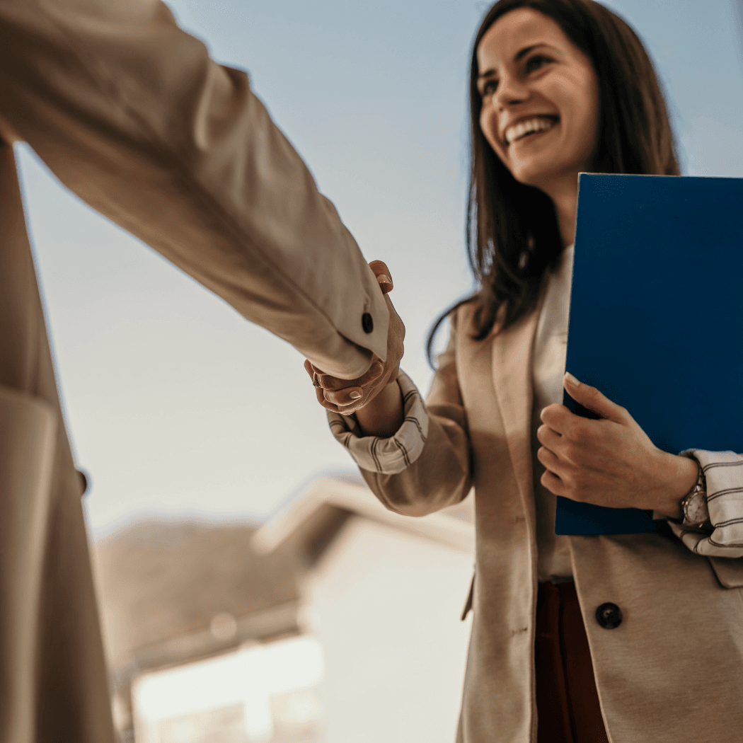 A smiling businesswoman holding a blue folder shakes hands with a client outdoors, viewed from a low angle.