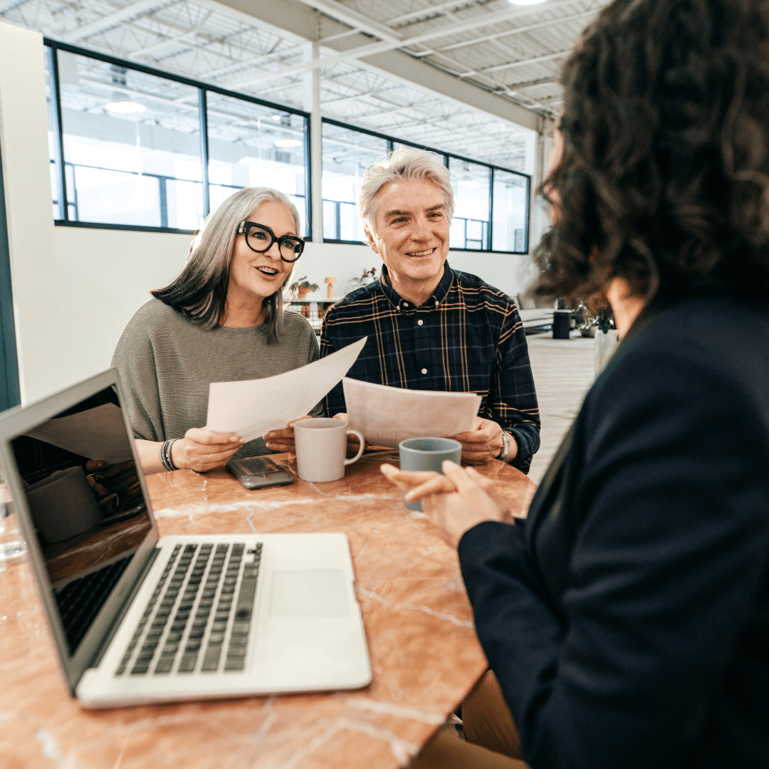 A smiling older couple with grey hair sits across from a financial advisor at a table, holding documents and coffee mugs during a consultation meeting.