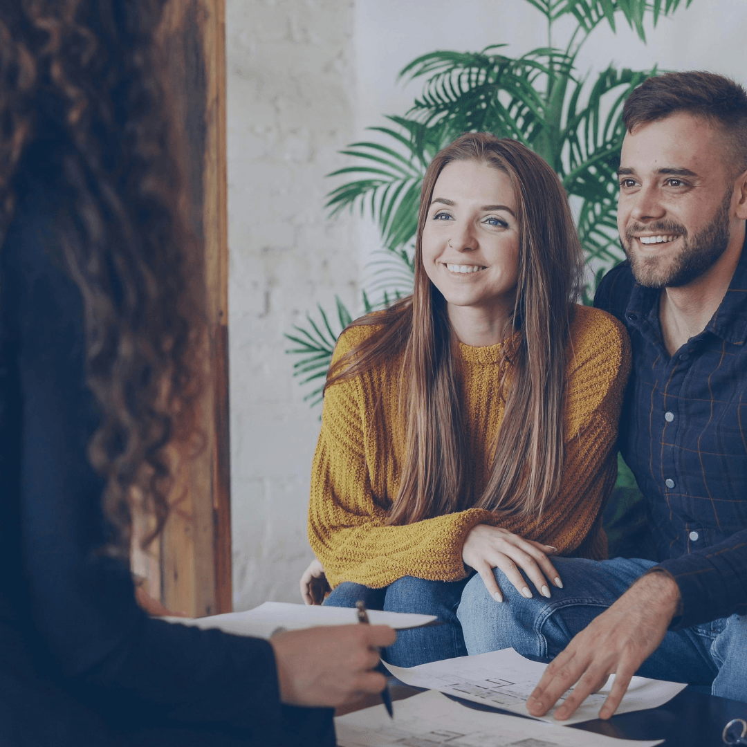 A young couple smiles attentively as a financial advisor reviews documents with them during a planning consultation in a warmly decorated office.