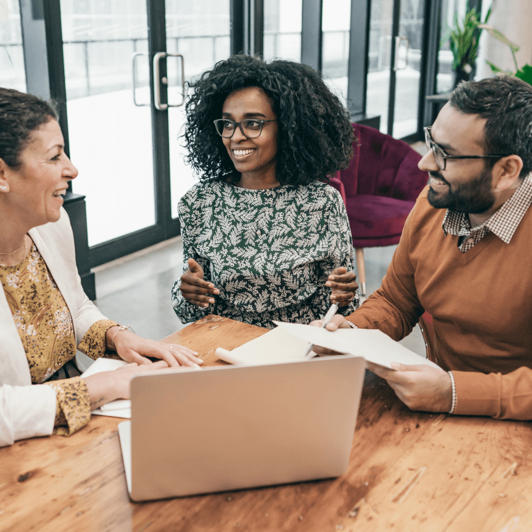Three colleagues gather around a laptop at a wooden table in a bright office, engaged in an animated and friendly business discussion.