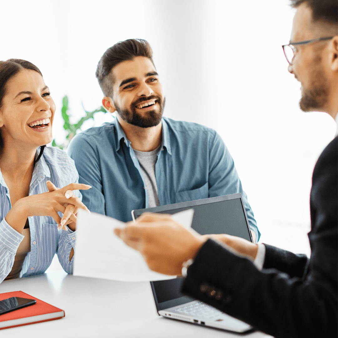 A laughing young couple sits across from a suited advisor who is handing them a document during a positive financial consultation meeting.