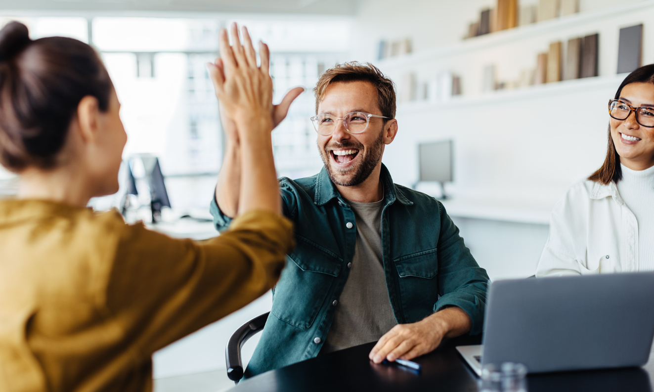 Happy coworkers high-fiving during a meeting while discussing group health insurance and mental health support.