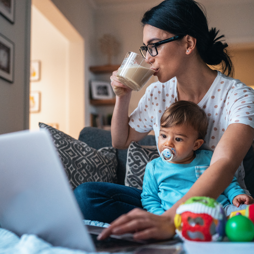 A mother wearing glasses drinks coffee while working on a laptop, with a toddler wearing a soother sitting on her lap.