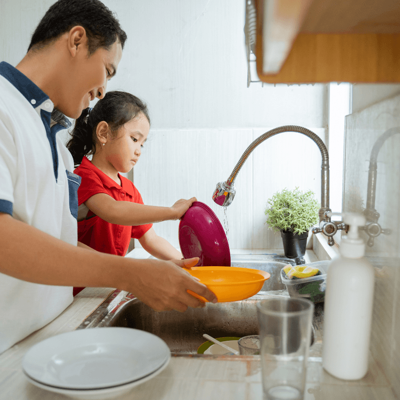 A father and young daughter wash dishes together at the kitchen sink.