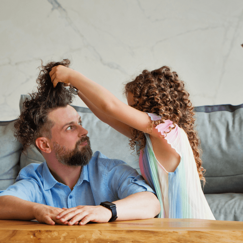 A young girl plays with her father's curly hair while he rests at a table.