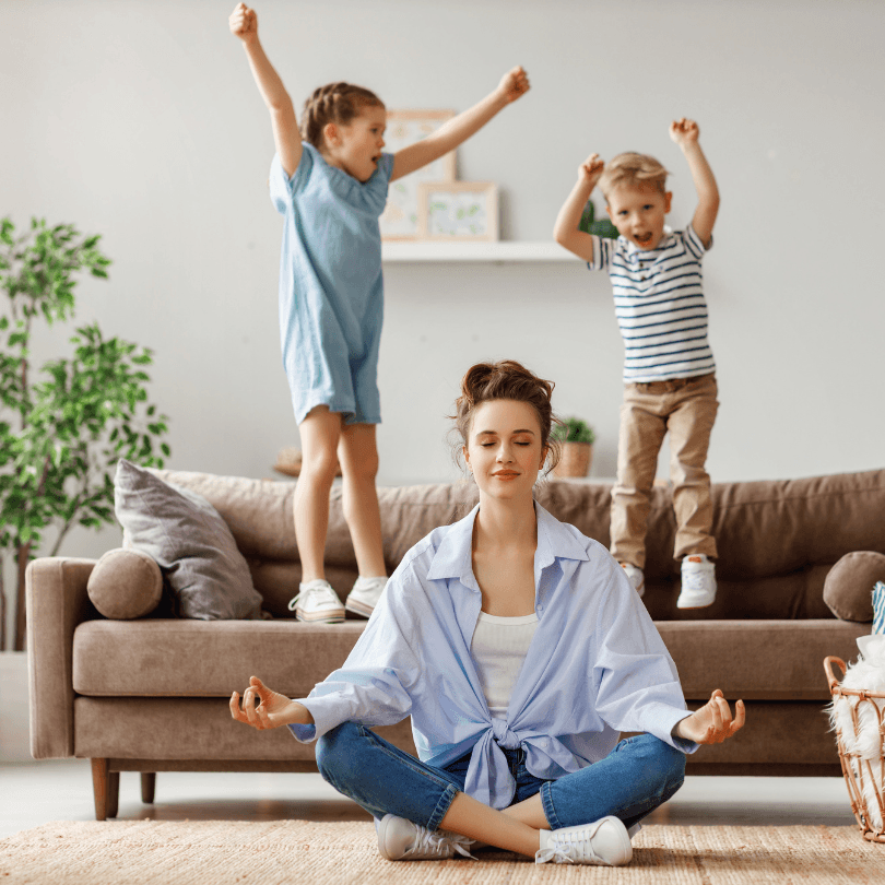 A mother sits cross-legged in a meditation pose while two children jump and cheer on the couch behind her.